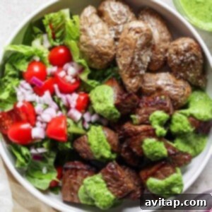 A vibrant bowl containing perfectly roasted potatoes, a fresh salad with crisp lettuce, juicy tomatoes, and red onions, topped with air fryer steak bites and a generous drizzle of green sauce.