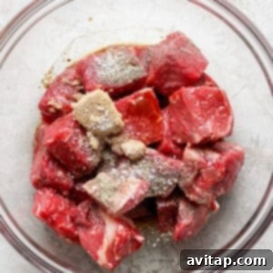 Effortless Air Fryer Steak Bites 9 A glass bowl filled with raw beef chunks seasoned with spices on top, placed on a light-colored textured surface.