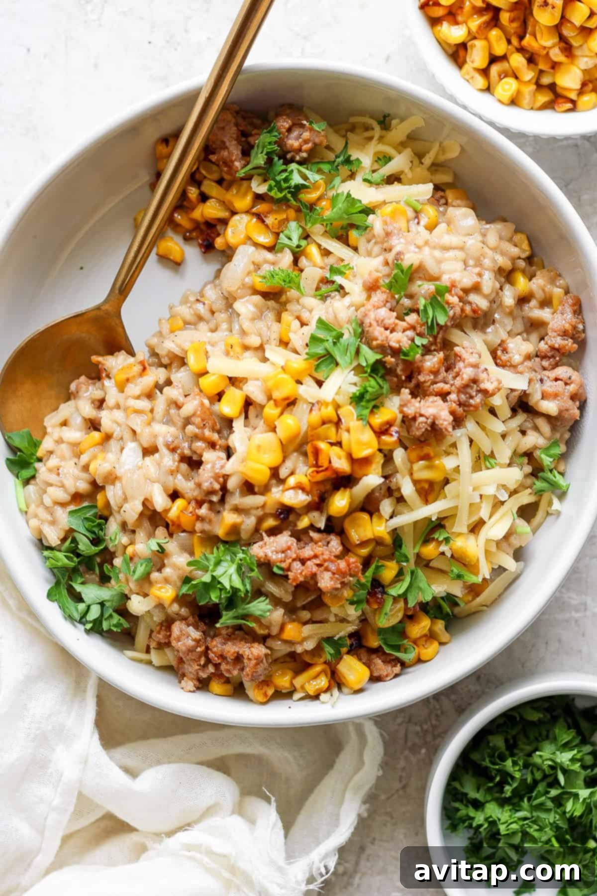 A beautifully presented bowl of creamy sweet corn risotto, generously topped with browned Italian sausage, vibrant corn kernels, a sprinkle of fresh parsley, and a hint of grated cheese. A elegant gold spoon rests beside the bowl, with a small dish of corn blurred in the background, hinting at the freshness of ingredients.