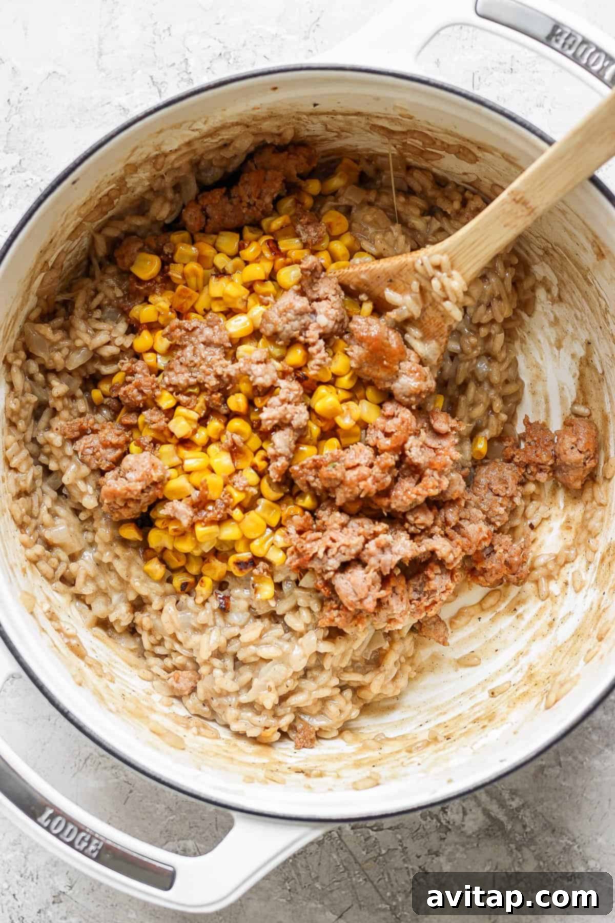 A close-up shot of a white Dutch oven on a textured surface, filled with creamy risotto. A wooden spoon is gently stirring the mixture, revealing golden corn kernels and pieces of ground sausage within the rich, off-white rice.
