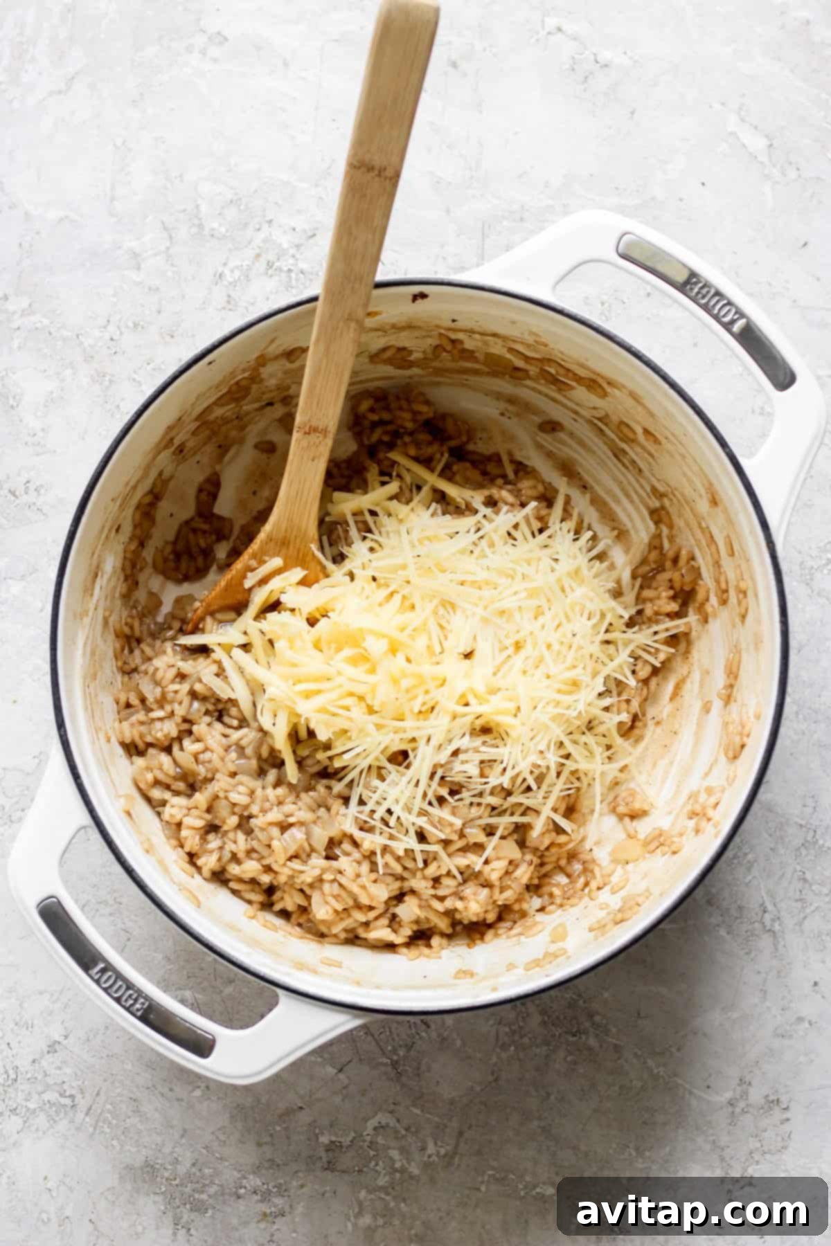 A close-up shot of a white Dutch oven on a light gray surface, with creamy risotto inside. A generous amount of grated cheese, a mix of white and yellow, is sprinkled over the top, ready to be stirred into the hot rice with a wooden spoon that rests in the pot.