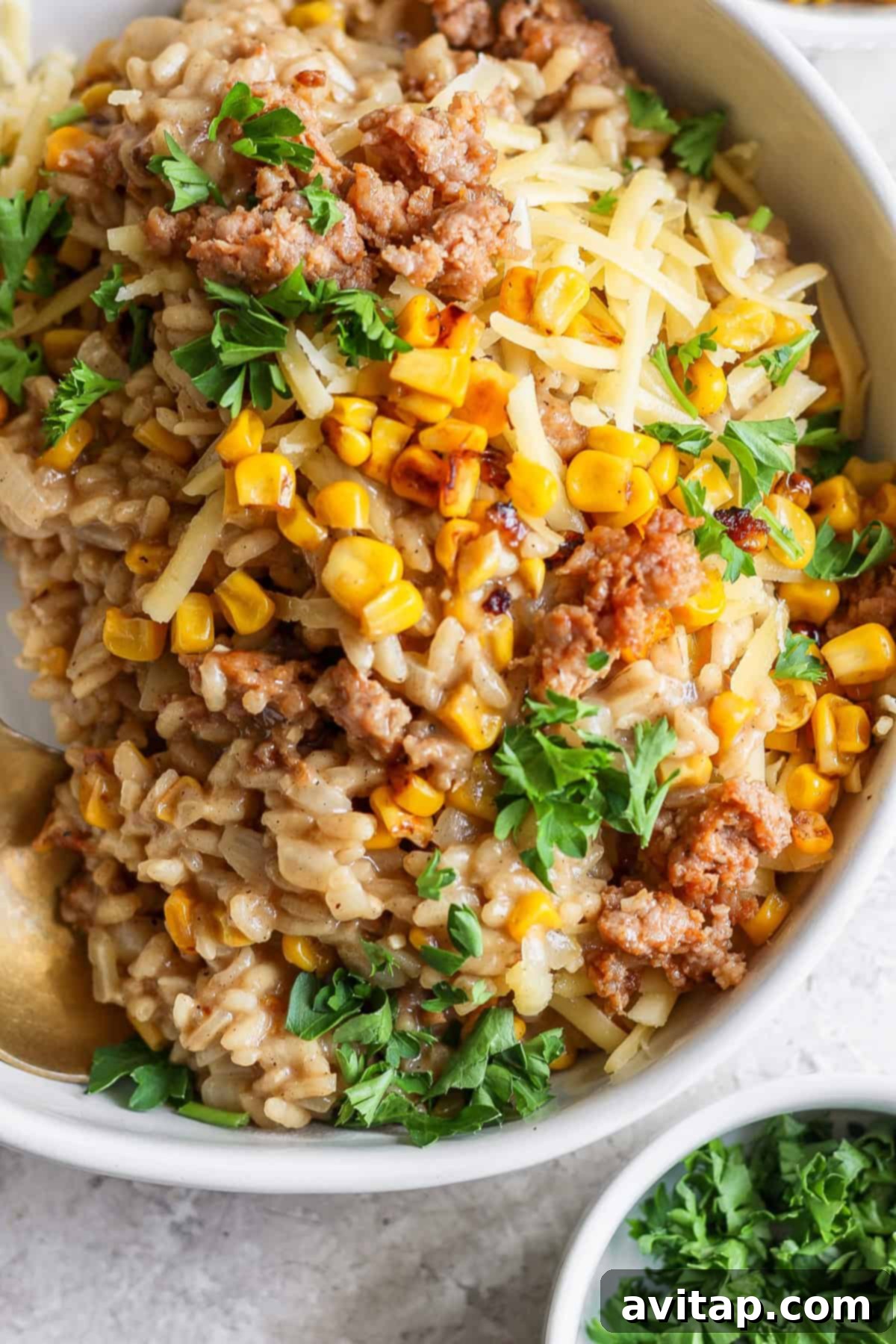 A close-up view of a beautifully prepared bowl of sweet corn risotto, adorned with perfectly browned ground meat, vibrant yellow corn kernels, a generous sprinkle of shredded cheese, and a garnish of fresh green parsley. In the soft-focus background, a small white dish holds additional chopped parsley, emphasizing freshness.