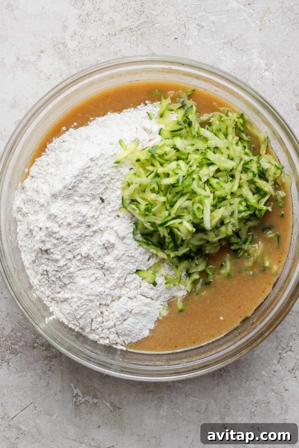 A glass bowl containing a baking mixture with flour, brown liquid batter, and grated zucchini on a light gray surface, highlighting the process of combining ingredients for the healthy zucchini cake.