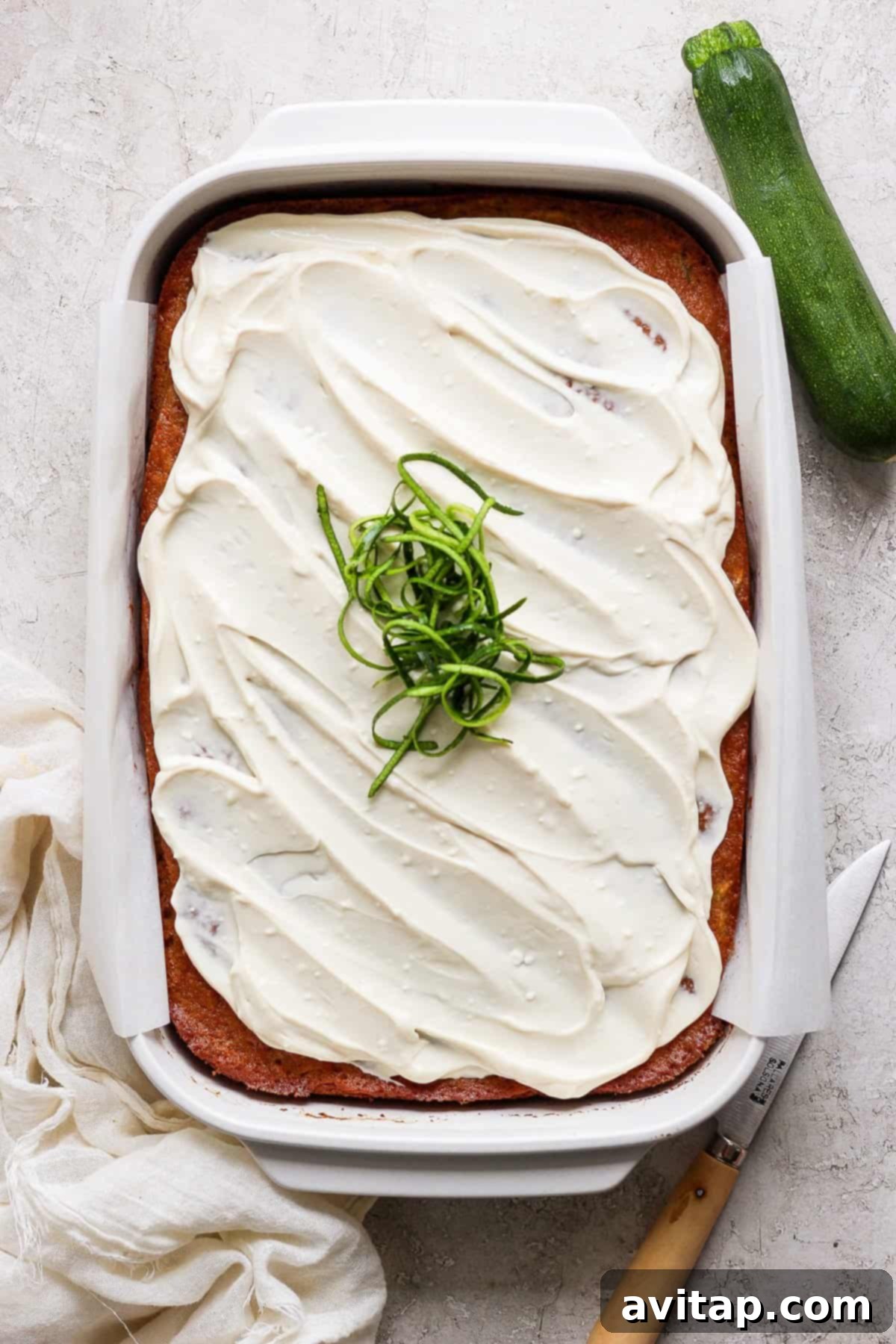 A freshly baked and frosted sheet cake, garnished with delicate spiralized zucchini ribbons, sits invitingly in a rectangular baking dish. A knife and a whole zucchini are placed nearby, suggesting preparation and serving.