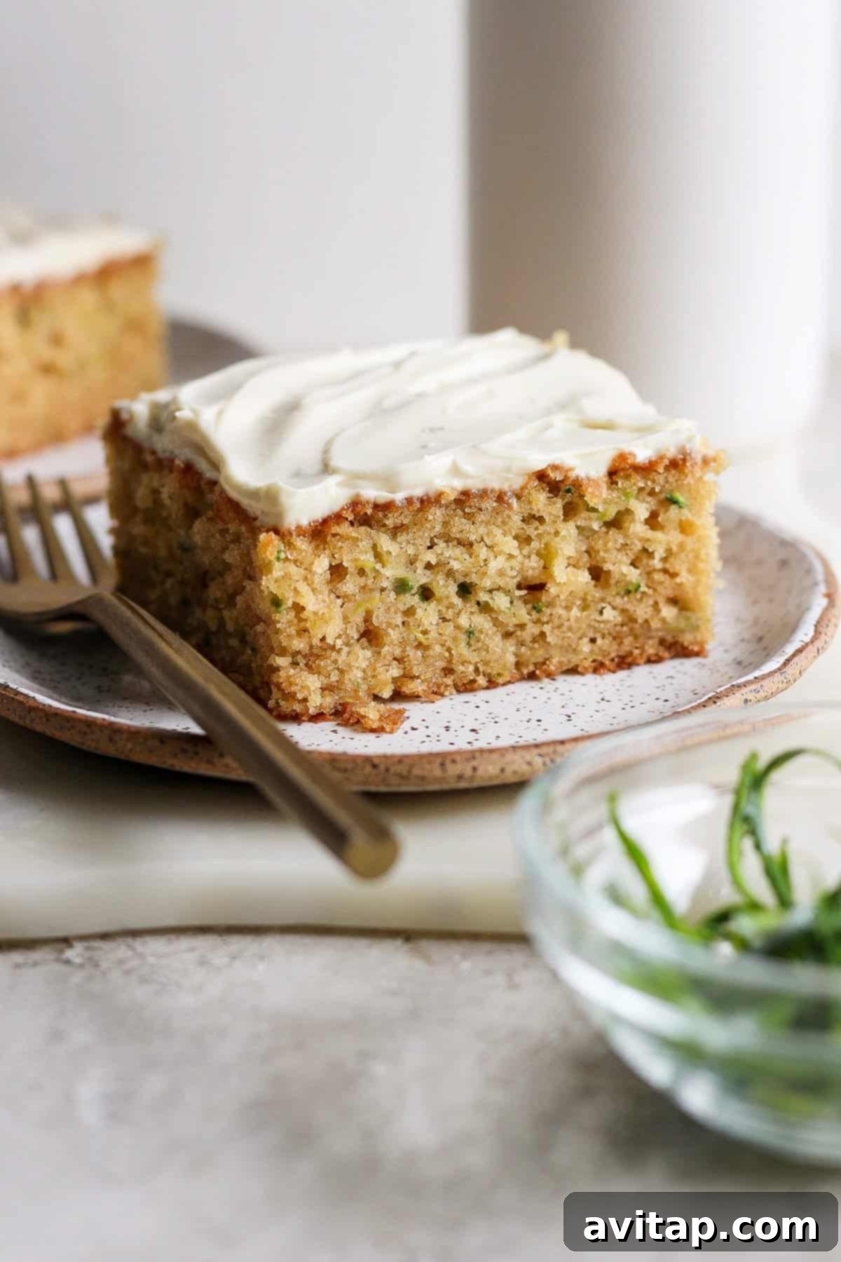 A perfectly frosted slice of healthy zucchini cake sits on a clean white plate, accompanied by a silver fork, with a bowl of vibrant green garnish blurred in the foreground, ready to be enjoyed.