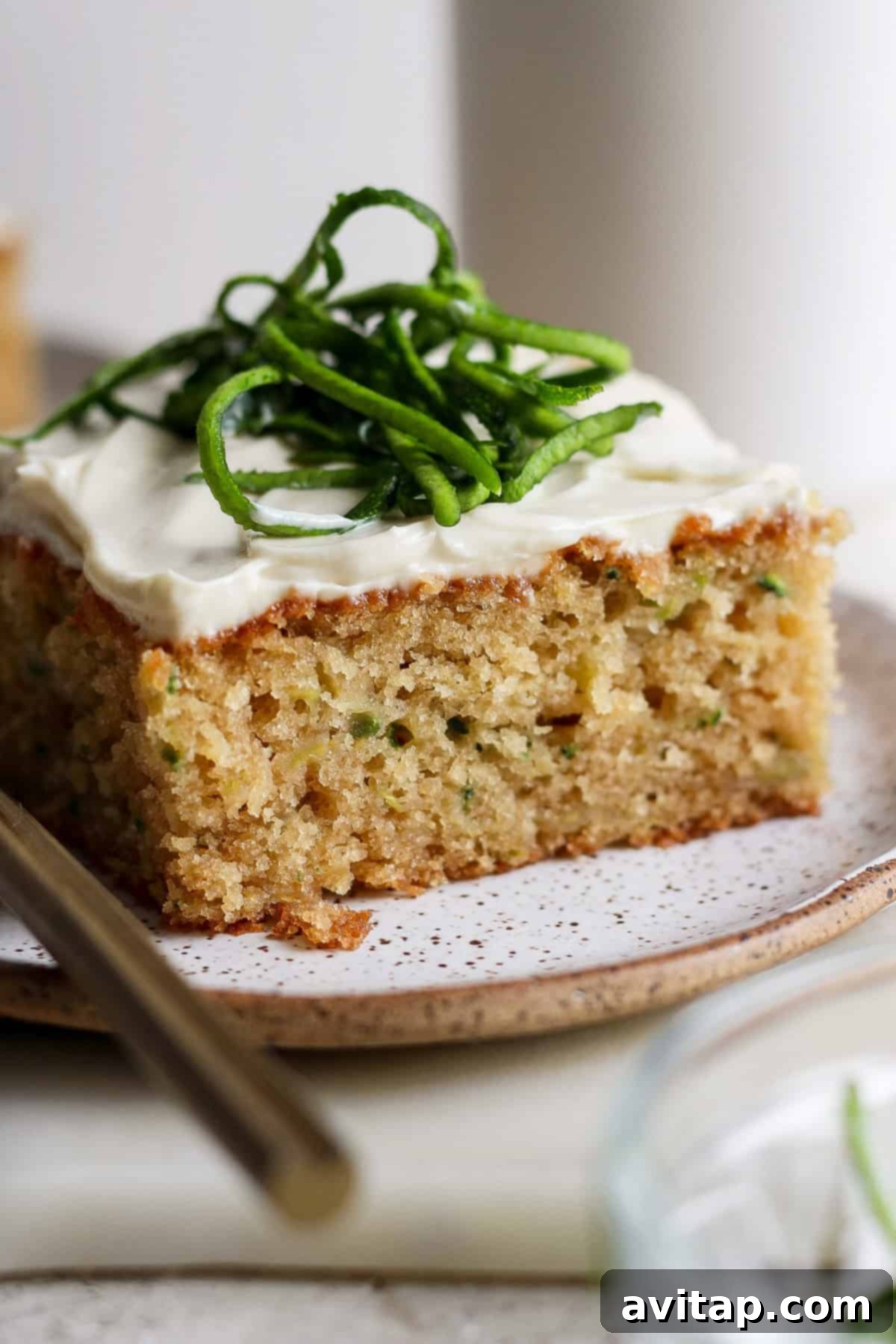 A single slice of moist zucchini cake, elegantly frosted with white cream cheese and adorned with delicate green zest, rests on a speckled plate with a fork beside it, inviting a delicious bite.
