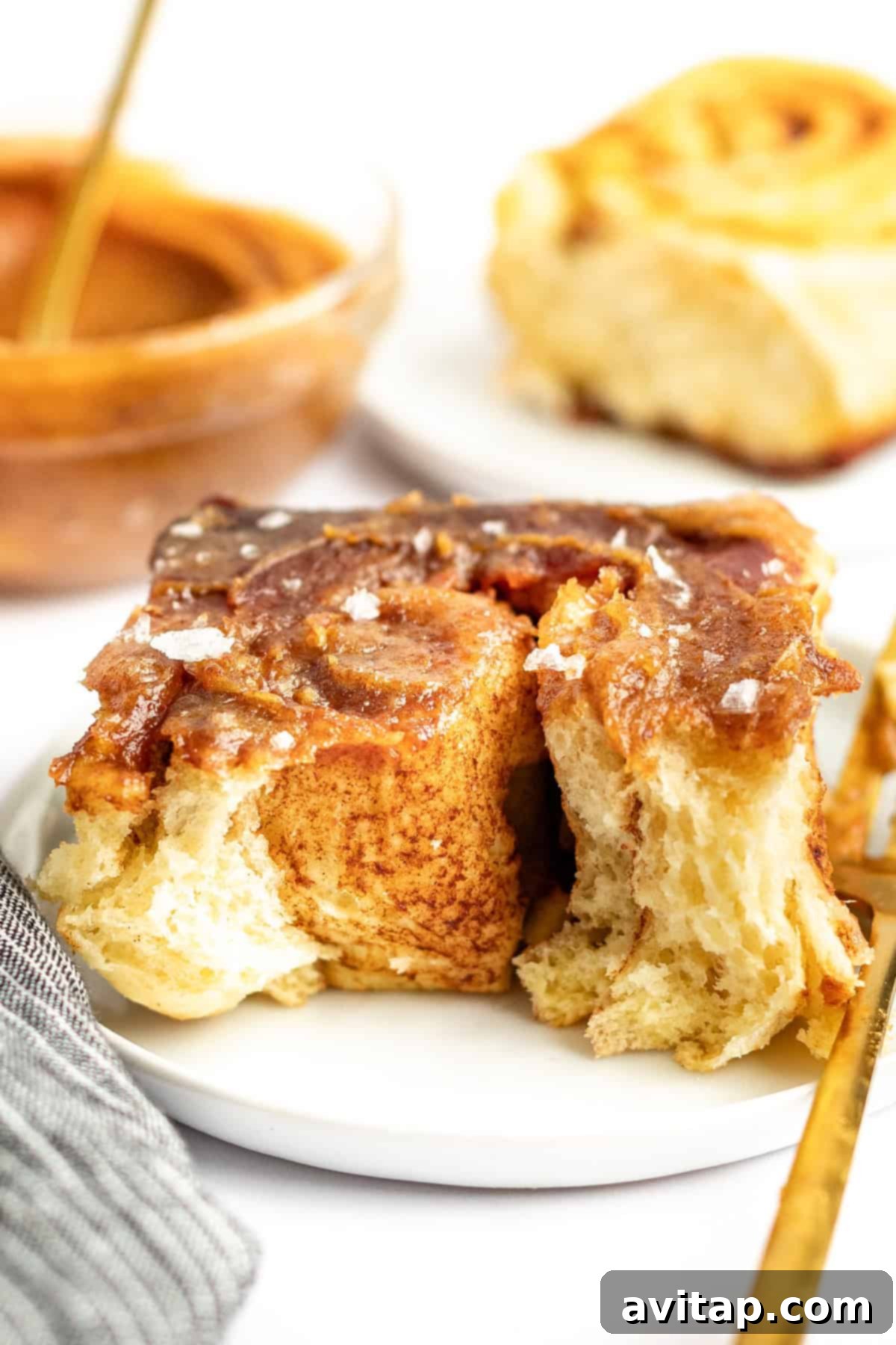 Close-up of a cinnamon roll with glaze and a sprinkle of salt on a white plate. A partially eaten cinnamon roll and a dish with more glaze are in the background.