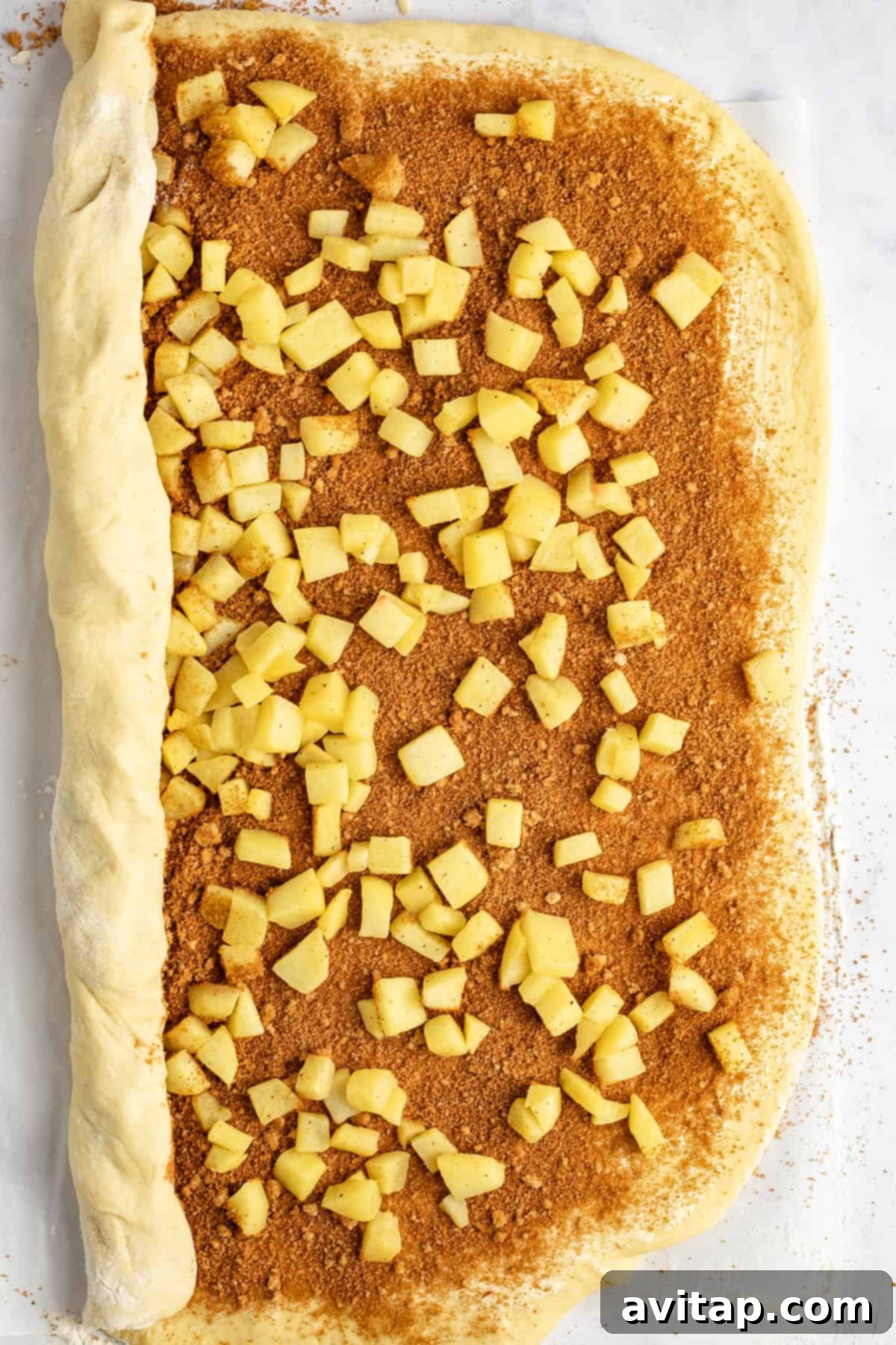 Unbaked cinnamon roll dough topped with diced apples and cinnamon sugar, partially rolled up on a light surface.