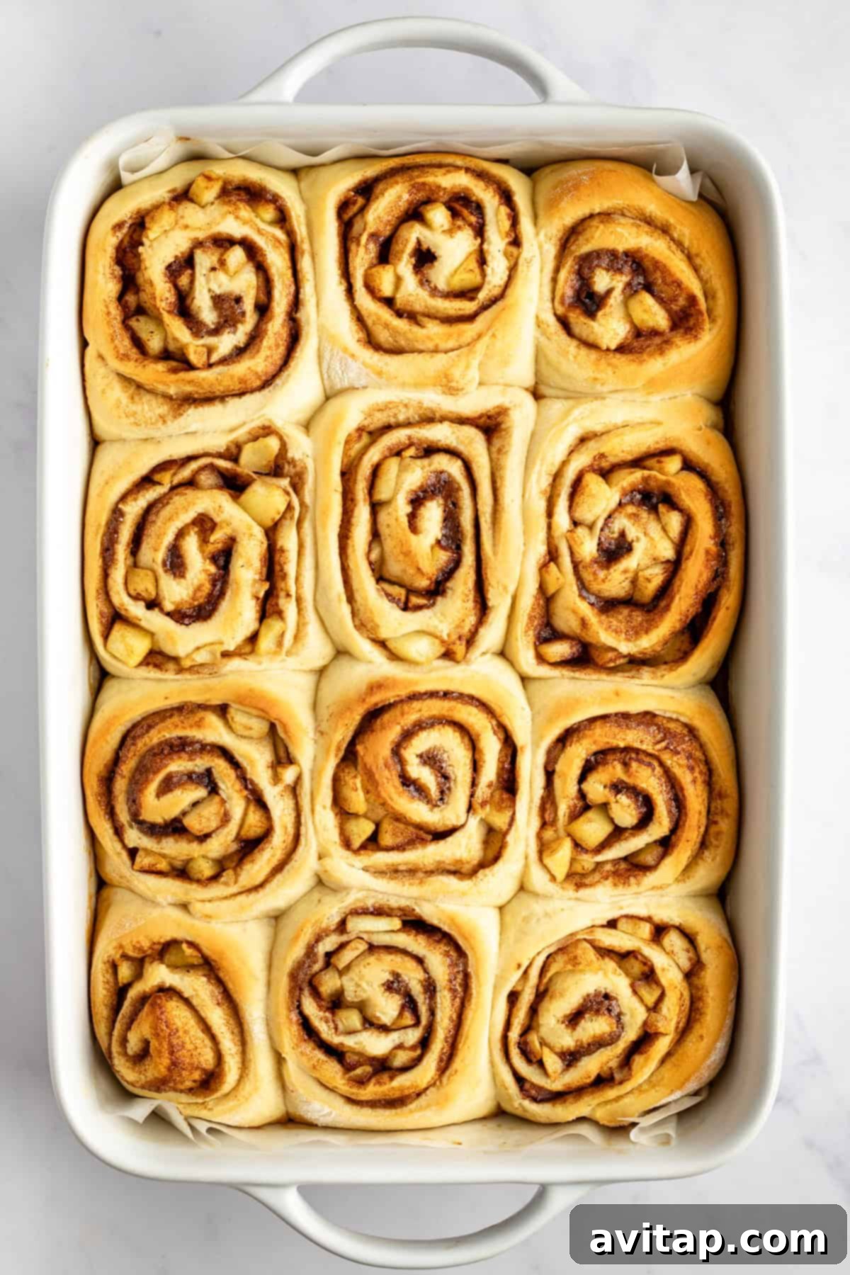 A rectangular baking dish filled with twelve freshly baked cinnamon rolls, each with a visible swirl pattern, on a white surface.