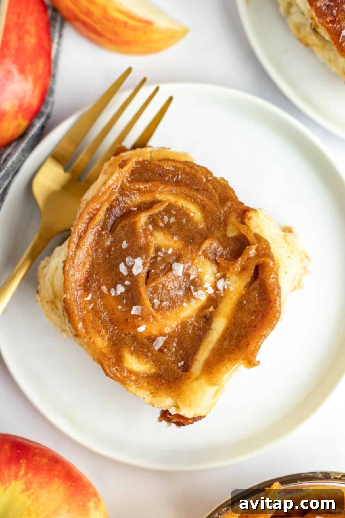 A single cinnamon roll with frosting and a sprinkle of sea salt is on a white plate with a gold fork. Slices of a red apple are partially visible in the background.