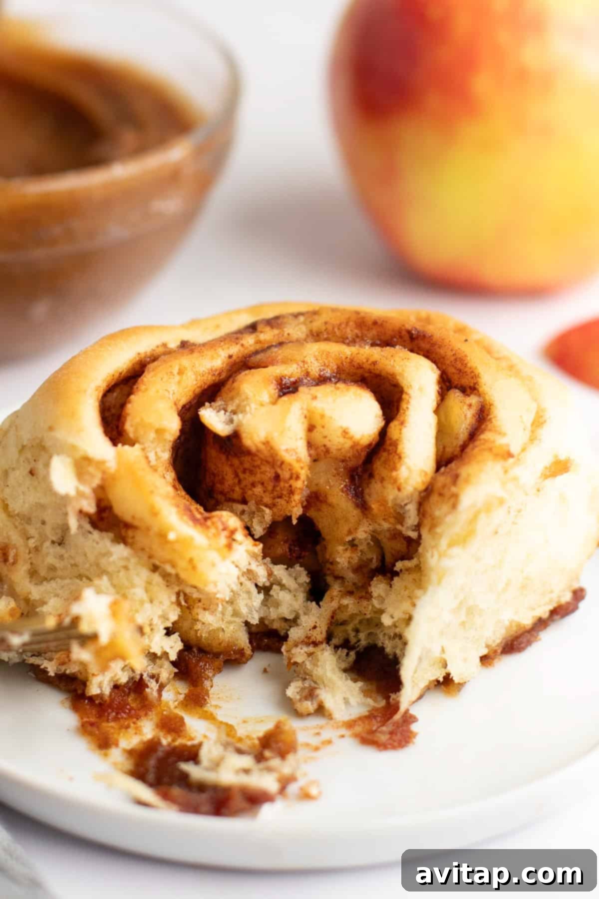 A partially eaten cinnamon roll on a white plate with a bowl of filling and an apple in the background.