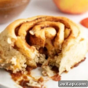 A close-up view of a partially eaten cinnamon roll on a white plate, showing its swirled layers and a bite taken out, with blurred background elements.