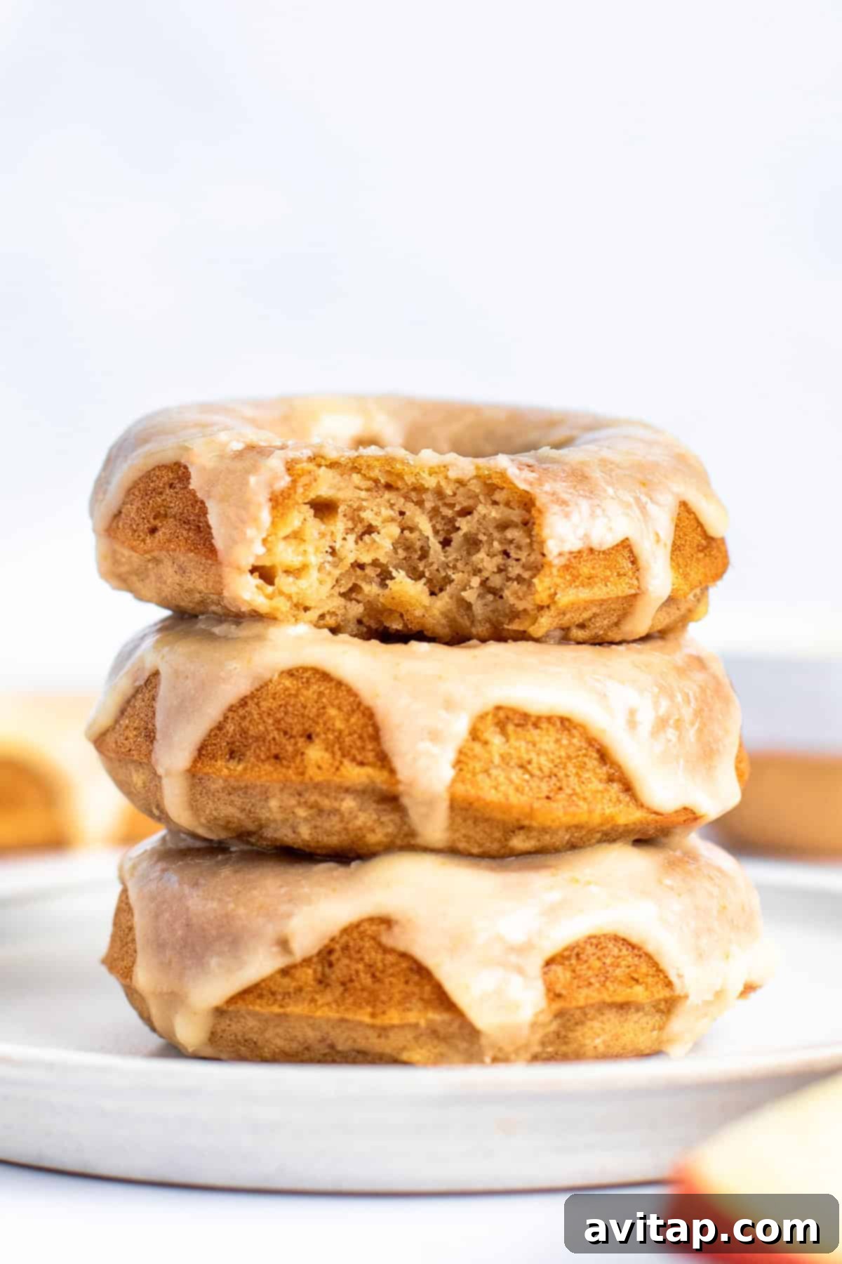 A stack of three glazed donuts on a white plate, with the top donut partially eaten, showing a golden brown interior.
