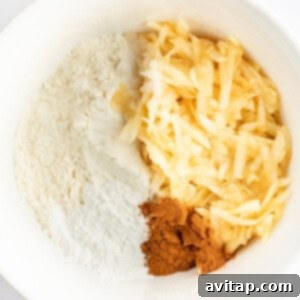 A white mixing bowl containing flour, grated apples, and a portion of ground cinnamon.