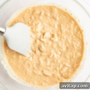 A glass bowl containing a thick, beige batter mixture with a white spatula resting on the side.