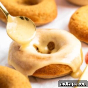 A spoon drizzles glaze over a baked donut with more donuts and a sliced apple in the background.