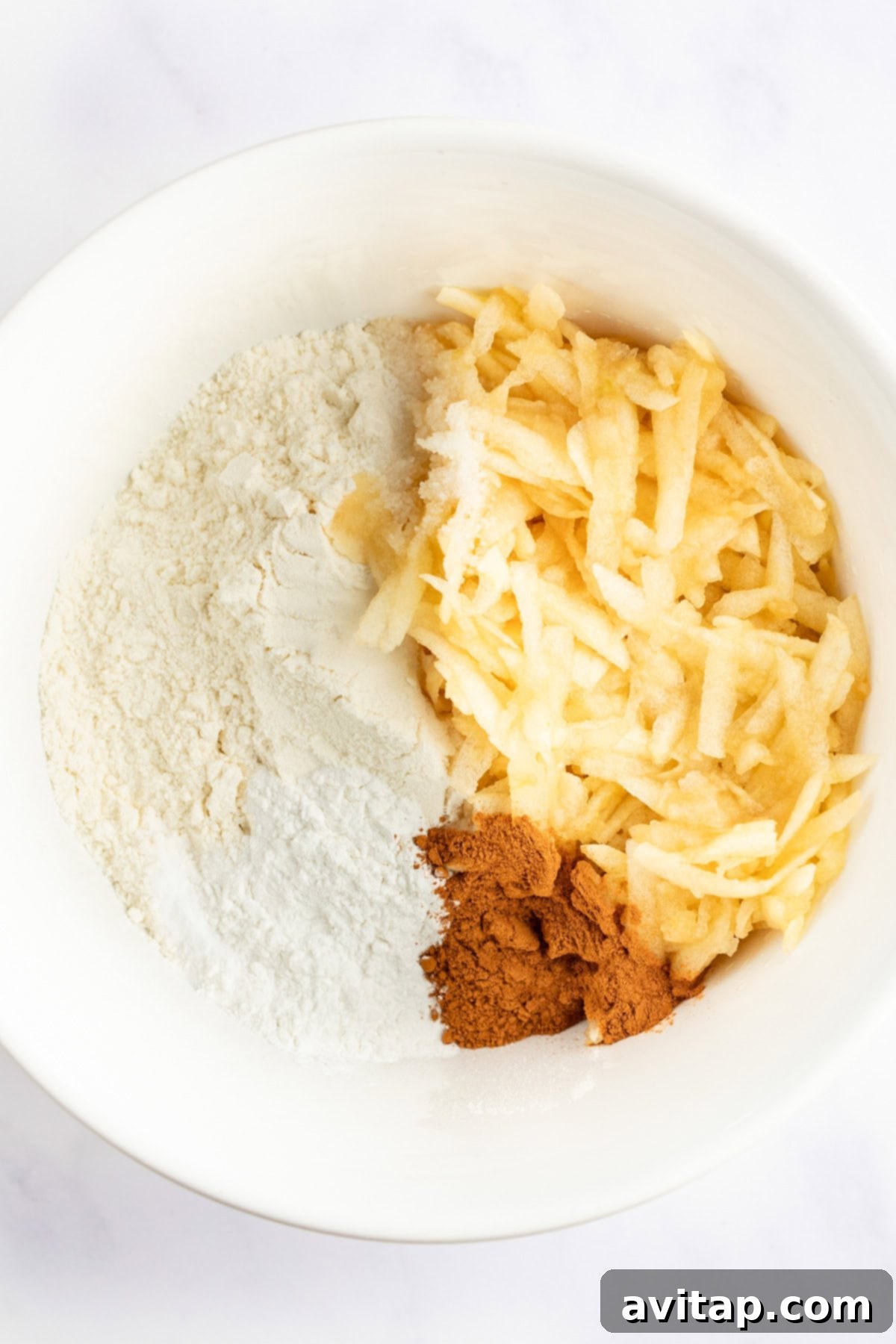 A white mixing bowl containing flour, grated apples, and a portion of ground cinnamon.