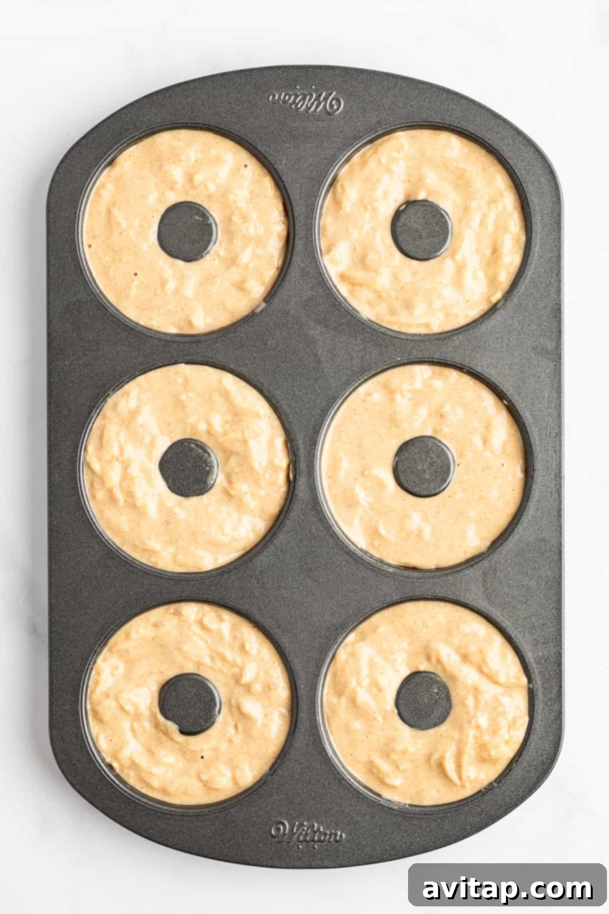 A six-cavity donut baking pan filled with unbaked donut batter, ready to be placed in the oven.