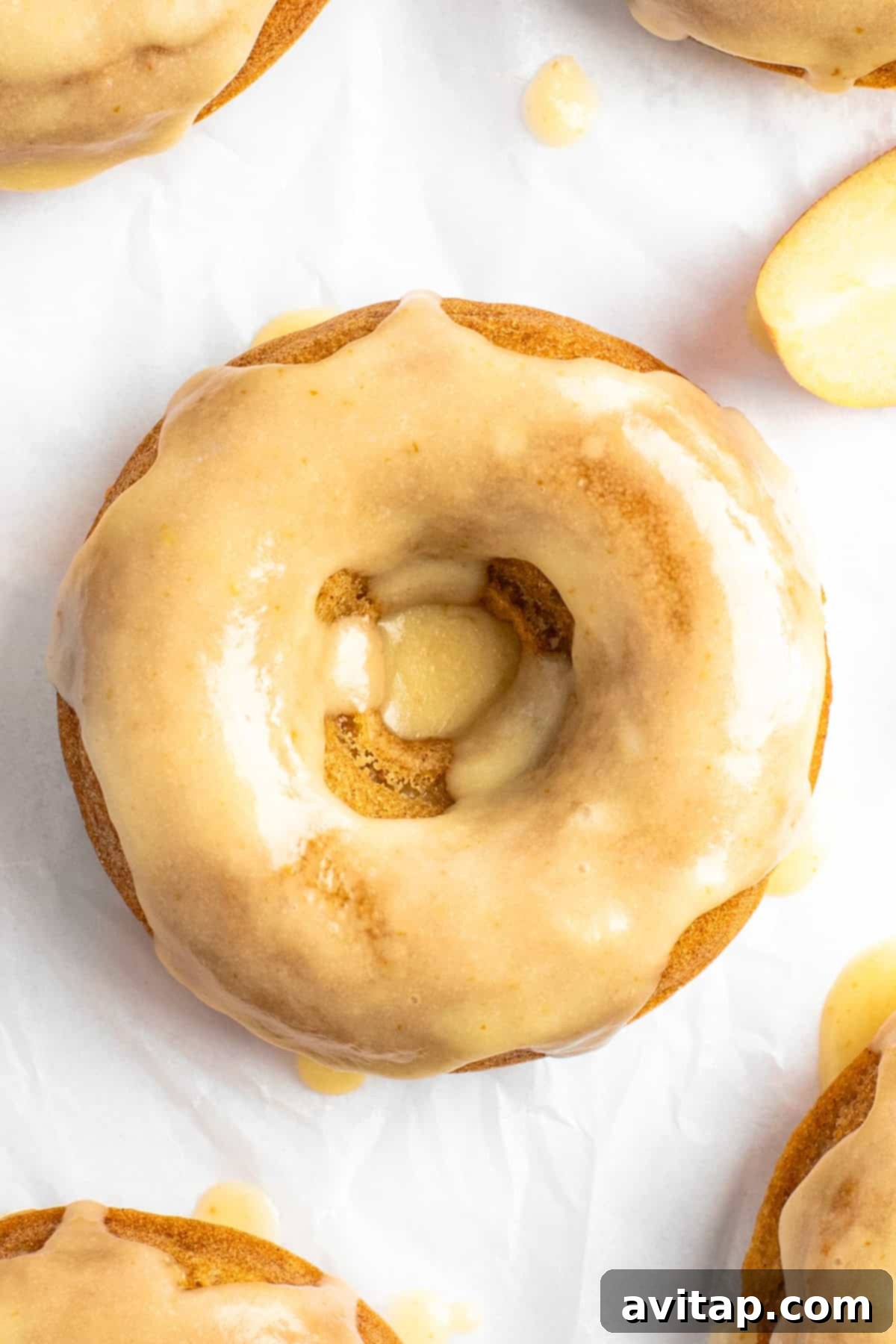 Close-up of a glazed donut with a smooth, shiny coating on a white surface. The donut has a hole in the center with some exposed texture. Another donut is partially visible in the background.