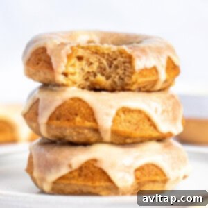 Three glazed donuts stacked on a plate, with the top donut having a bite taken out of it. The glaze is dripping down the sides of the donuts. The background is out of focus.