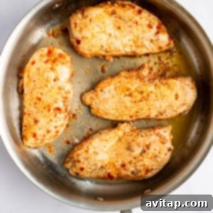 Four seasoned chicken breasts searing to a golden brown in a stainless steel skillet.