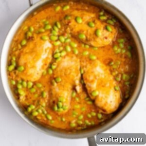 Cooked chicken breasts in a golden sauce with green edamame simmering in a stainless steel pan.