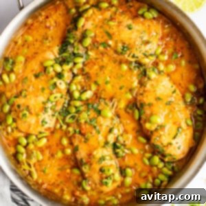 A stainless steel pan filled with cooked chicken in a vibrant orange sauce, garnished with chopped herbs and edamame beans. Lime wedges and a striped kitchen towel are placed beside the pan.
