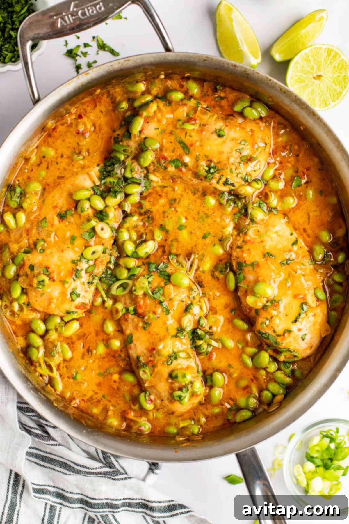 A large stainless steel pan showcasing beautifully cooked chicken fillets bathed in a rich, vibrant orange sauce, generously garnished with fresh herbs and bright green edamame beans. Fresh lime wedges and a stylish striped kitchen towel rest nearby.