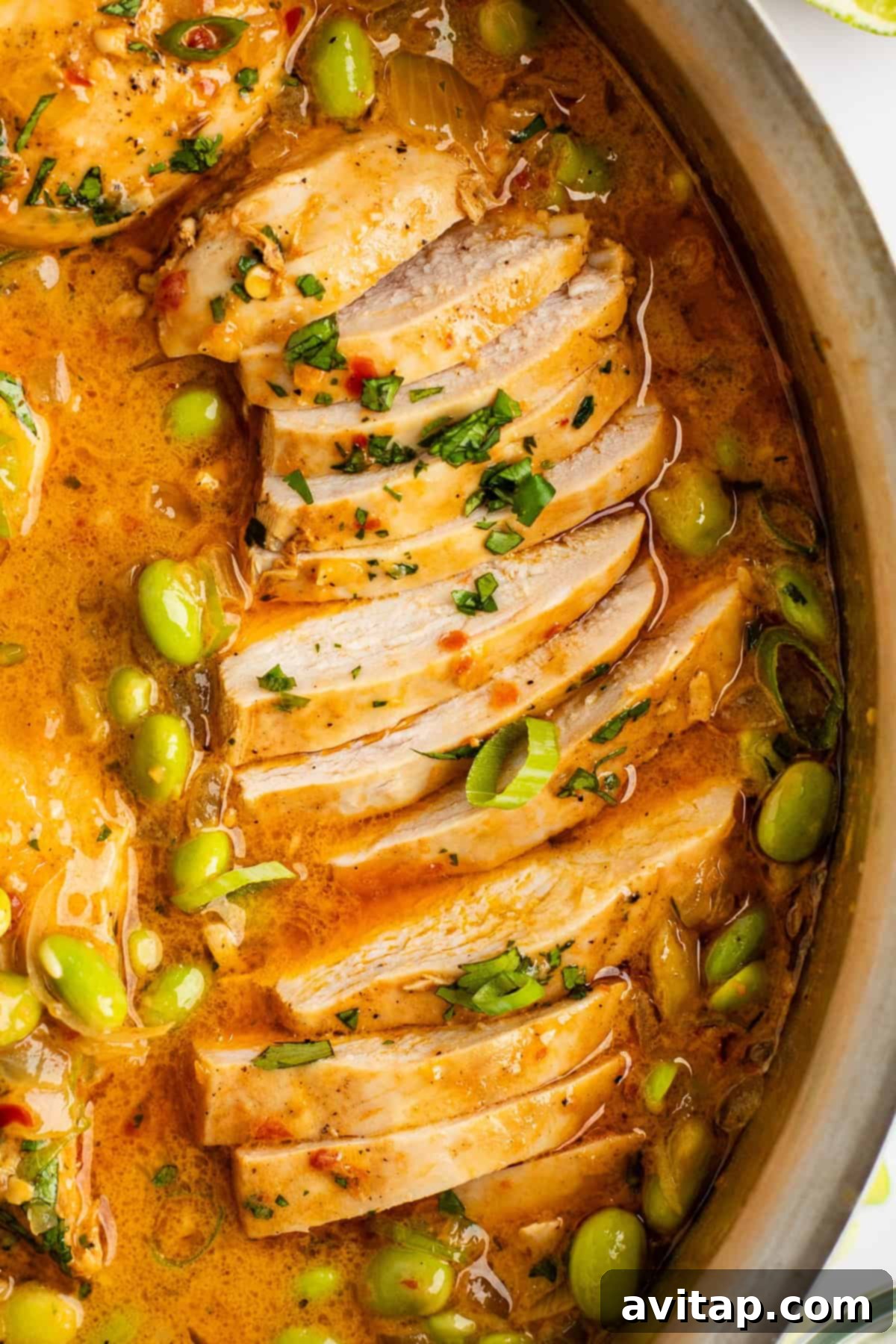 A close-up view of perfectly sliced, tender cooked chicken breast resting in a creamy, golden-orange sauce, beautifully garnished with finely chopped fresh herbs and bright green edamame peas, all within a rustic pan.