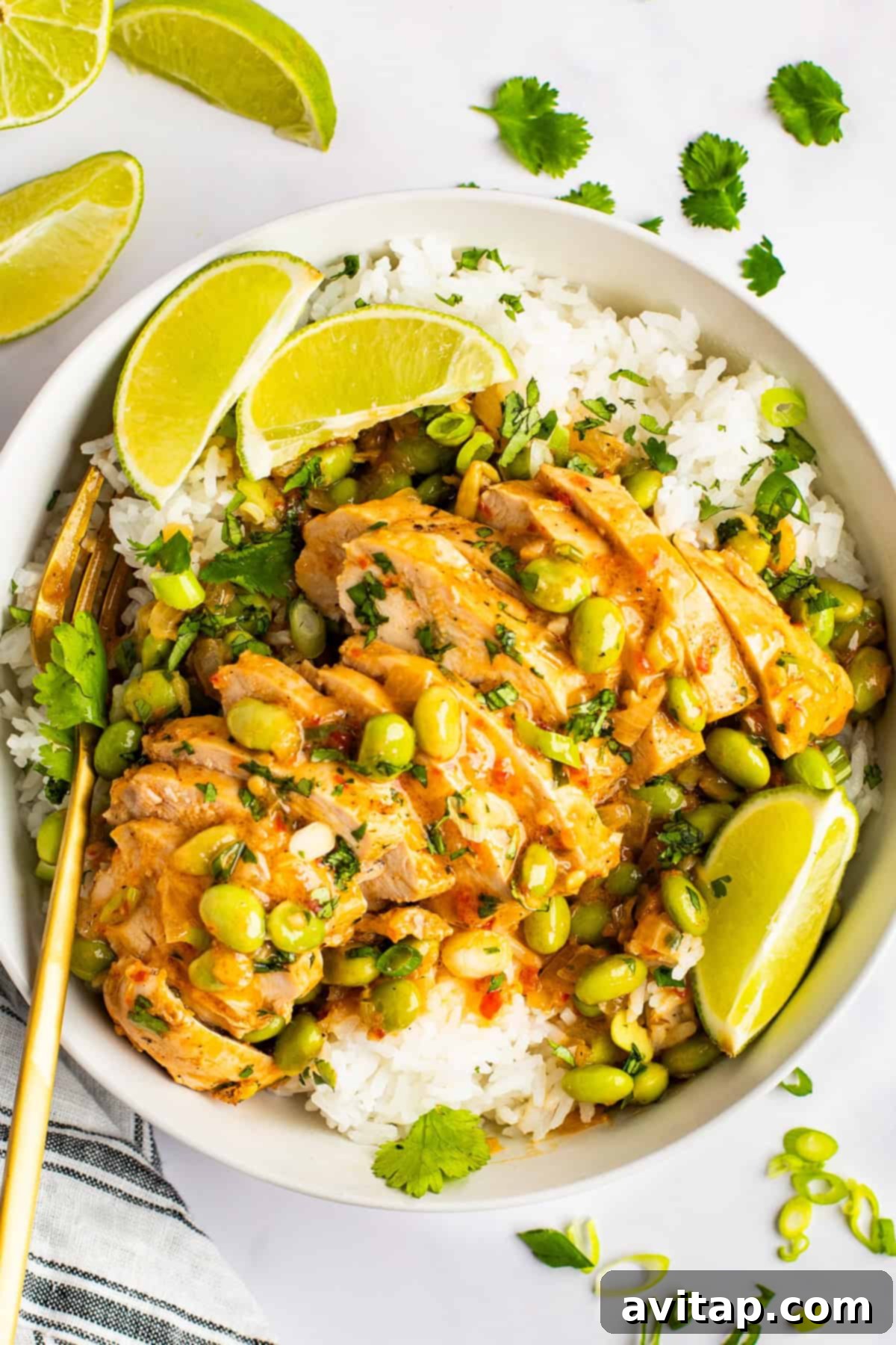 A beautifully composed bowl of Skillet Coconut Chicken served over fluffy white rice, adorned with bright green soybeans, a rich creamy sauce, fresh lime wedges, and cilantro. Golden chopsticks rest elegantly beside the bowl, with scattered lime slices completing the presentation.