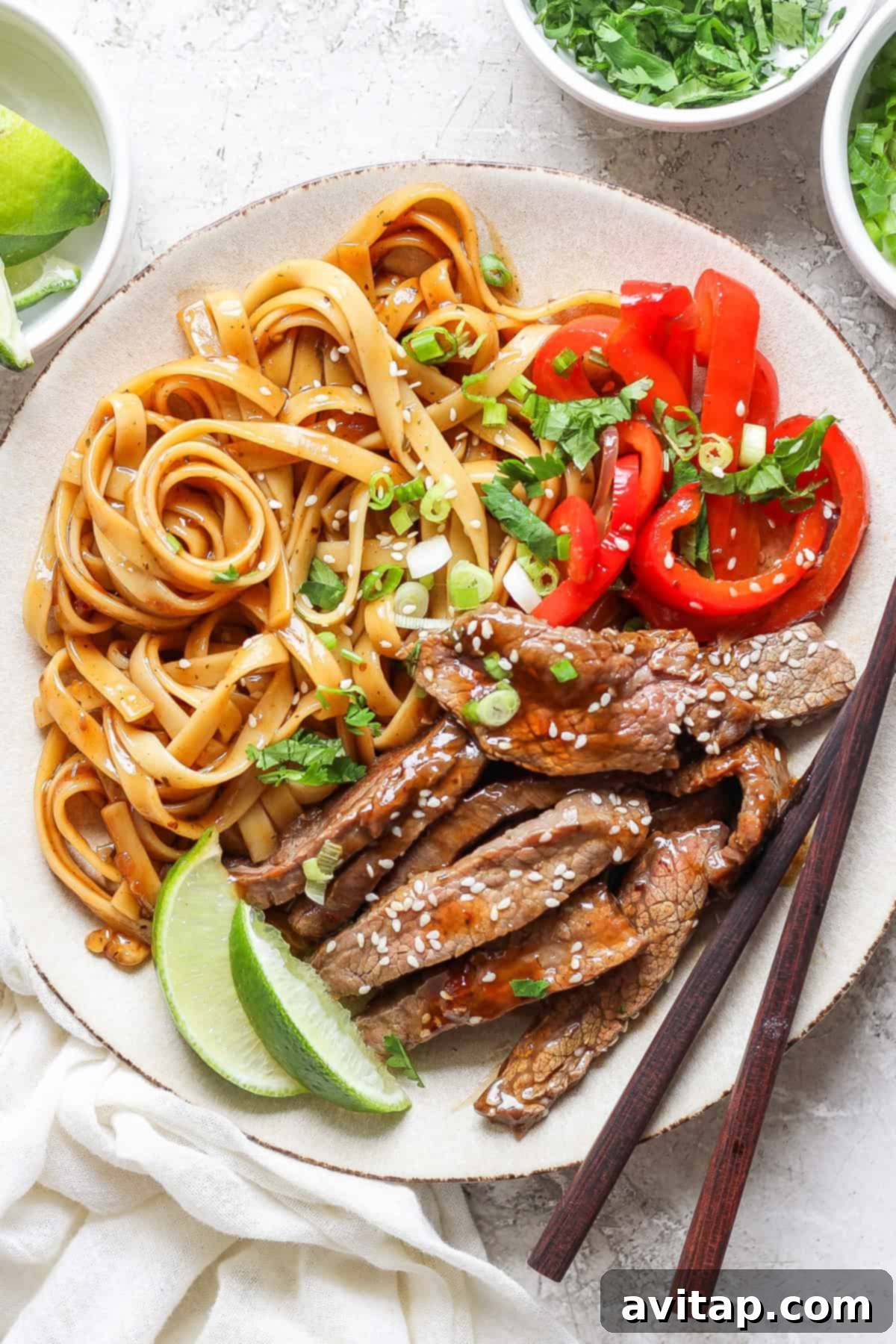 A plate with cooked noodles, sliced red bell peppers, and slices of beef topped with sesame seeds. A lime wedge is on the side, and a pair of chopsticks rests on the plate.