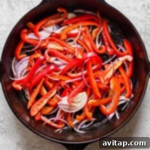 A cast iron skillet filled with sliced red bell peppers and red onions on a light-textured surface.