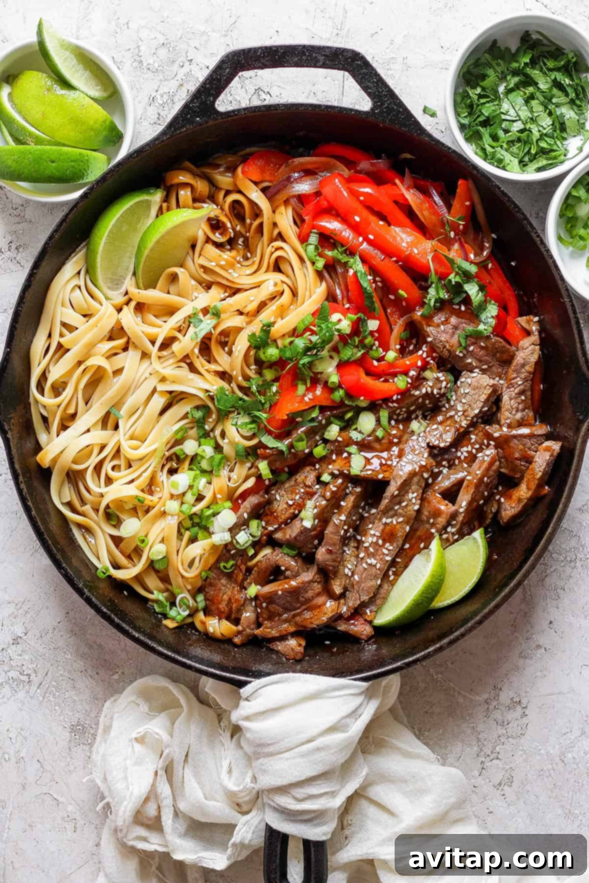 A cast iron skillet contains cooked noodles, sliced beef, red bell peppers, and lime wedges. Green herbs and lime slices are in small bowls nearby. A white cloth is draped over the skillet handle.