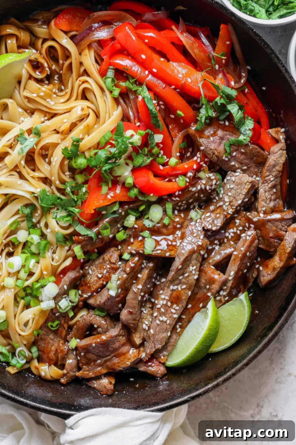 A bowl of stir-fried beef strips with sesame seeds, bell peppers, and noodles, garnished with chopped green onions and cilantro, alongside lime wedges.
