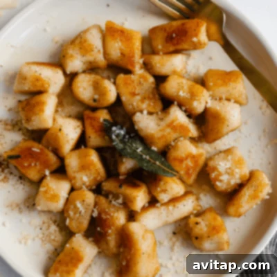 A plate of golden-brown cottage cheese gnocchi, beautifully adorned with crispy fried sage leaves and a generous sprinkle of grated cheese, accompanied by an elegant gold fork.