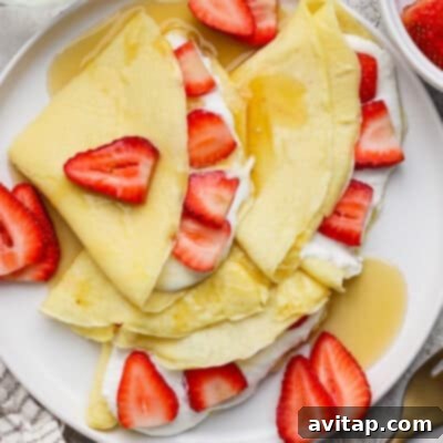 Delicate crepes, folded and artfully topped with fresh strawberry slices, a dollop of cream, and a generous drizzle of golden syrup on a pristine white plate.