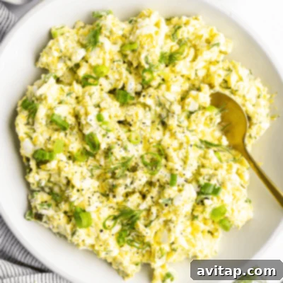 A bowl of creamy egg salad, generously garnished with fresh chopped green onions and fragrant dill, accompanied by an elegant gold spoon.