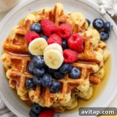 A tall stack of golden-brown cottage cheese waffles, generously topped with an assortment of fresh blueberries, raspberries, and banana slices, elegantly drizzled with sweet syrup on a pristine round white plate.