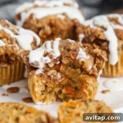 A close-up view of two delectable carrot muffins, one with a bite taken to reveal its moist interior dotted with carrot pieces. Both are crowned with a crumbly streusel topping and a delicate white icing drizzle.