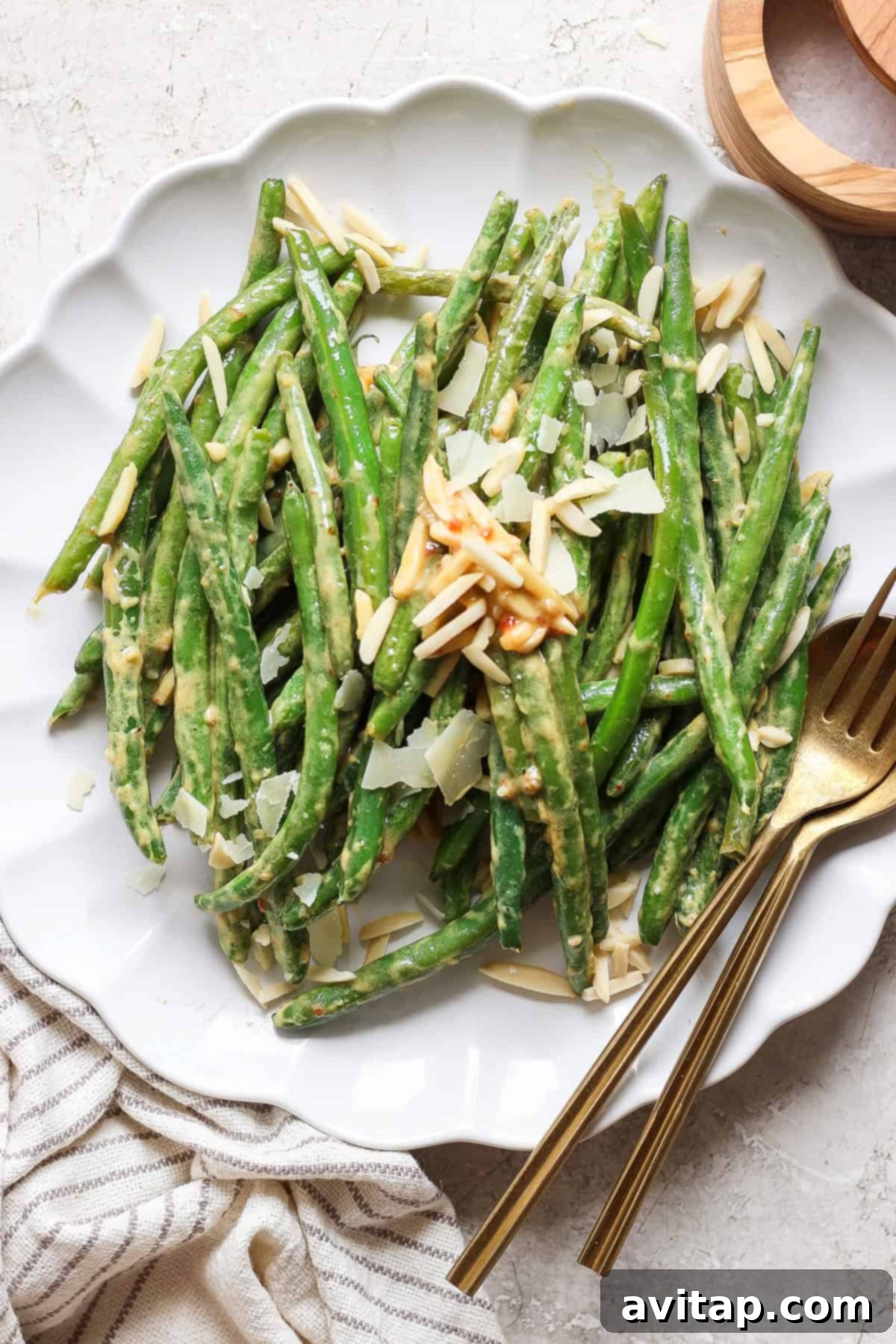 A plate of perfectly cooked green beans, elegantly topped with crunchy sliced almonds and a generous sprinkle of shaved parmesan cheese, served with a gleaming gold fork and knife on a crisp white tablecloth.