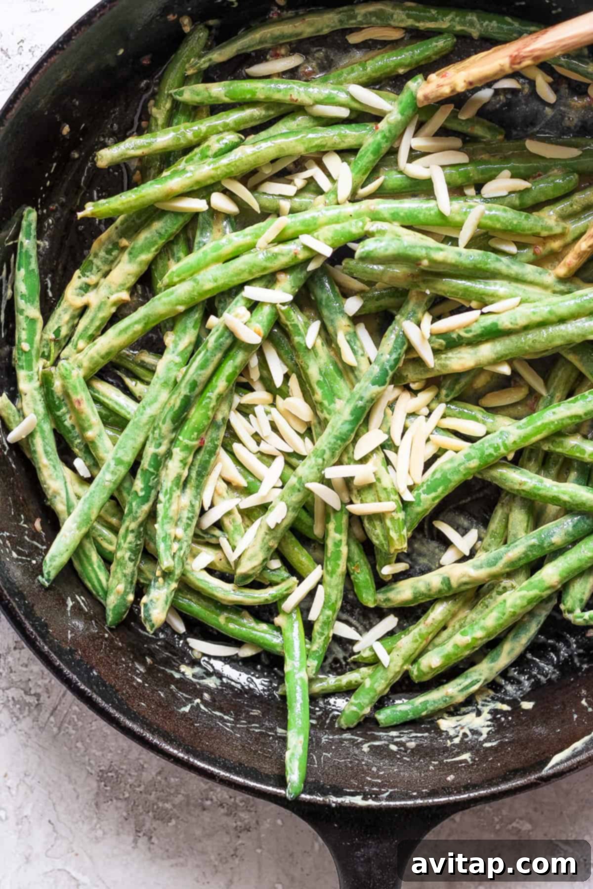 A rustic cast iron skillet brimming with vibrant green beans, glistening with a creamy, savory sauce, and generously crowned with golden slivered almonds. A wooden spoon rests invitingly in the upper right corner, ready to serve.