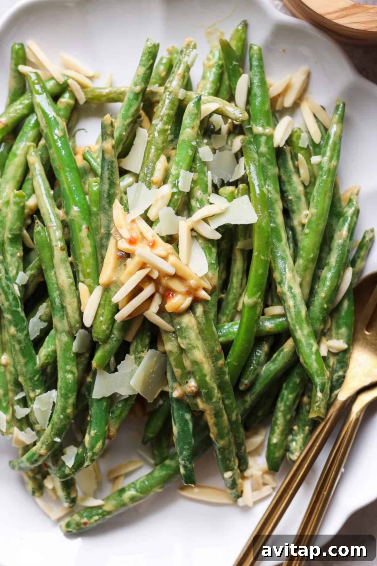 A beautifully presented plate of vibrant green beans, artfully adorned with delicate shaved Parmesan cheese and crunchy almond slivers. Elegant serving utensils are positioned neatly on the side, inviting you to enjoy this delicious and wholesome dish.