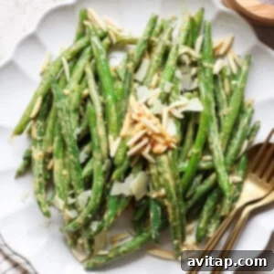 A plate of vibrant green beans topped with delicate shaved almonds and cheese, accompanied by a fork and knife, inviting you to savor its fresh taste.