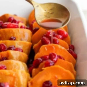 A spoon drizzles liquid over sliced sweet potatoes topped with cranberries in a rectangular white baking dish.