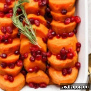 A white casserole dish filled with neatly arranged sliced sweet potatoes, topped with cranberries and garnished with a sprig of rosemary.