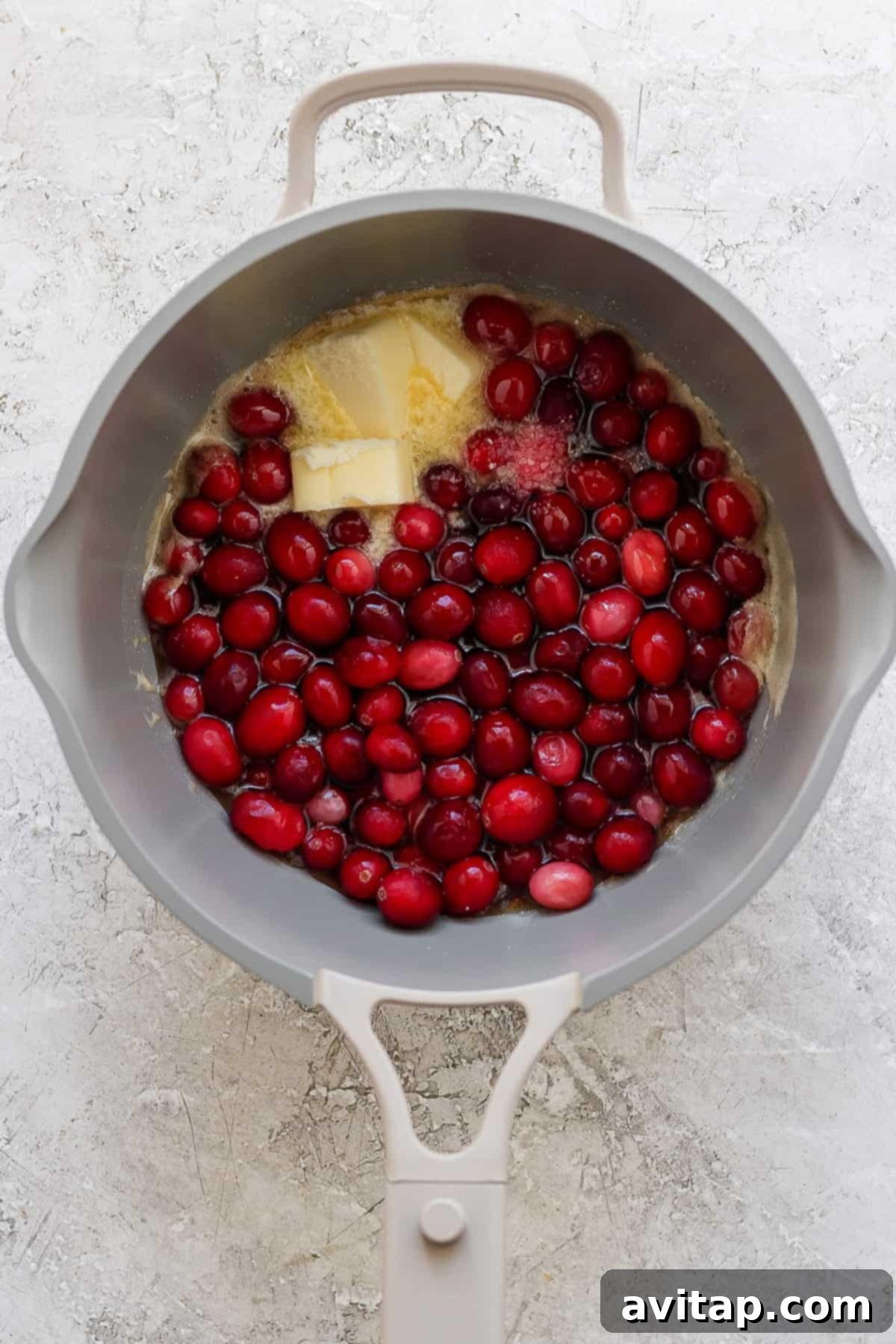 A gray pot contains raw cranberries, a block of butter, and sugar on a light textured surface.