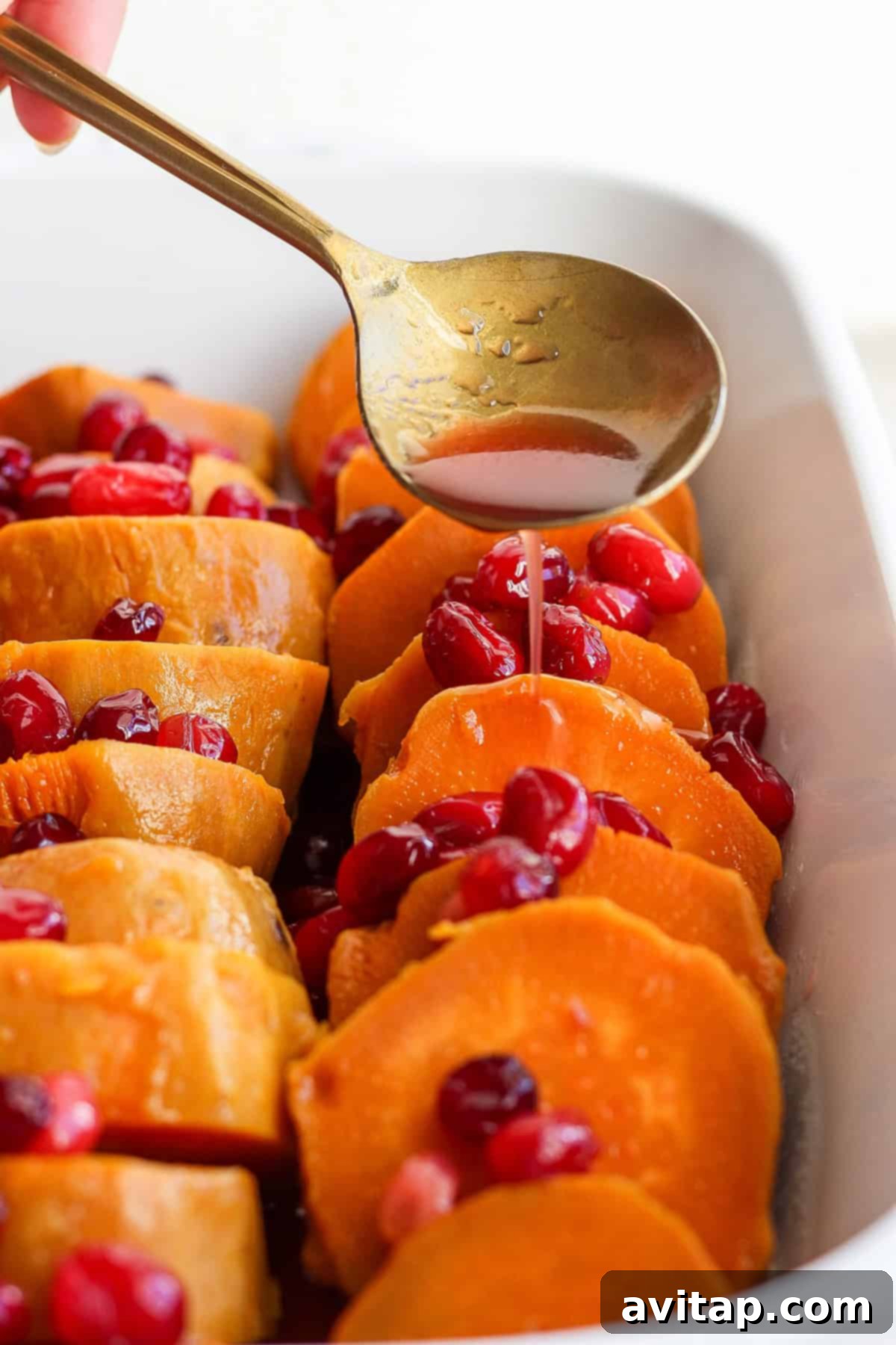A spoon drizzles liquid over sliced sweet potatoes topped with cranberries in a rectangular white baking dish.