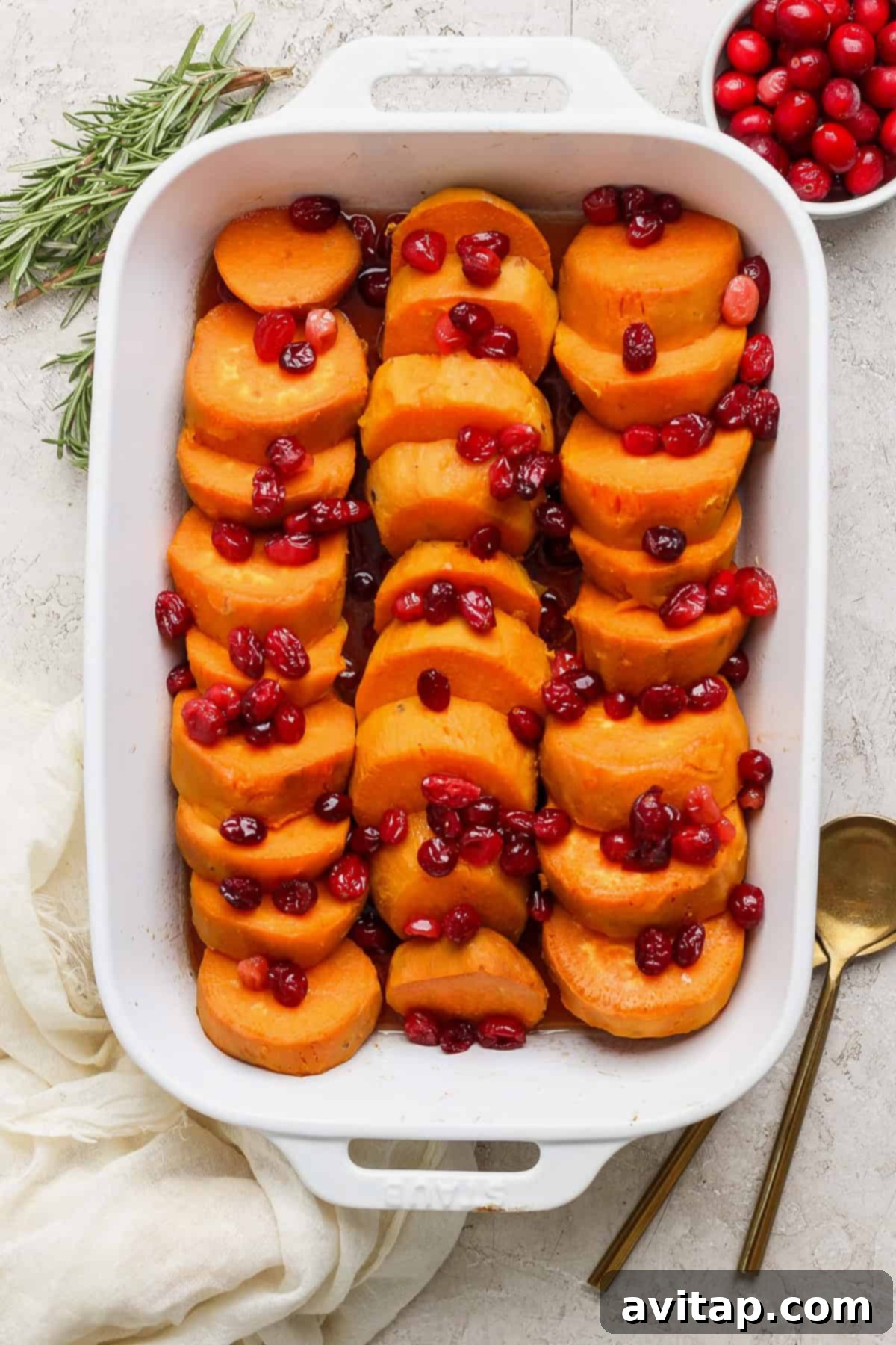 A white baking dish filled with sliced sweet potatoes topped with cranberries. A sprig of rosemary, a bowl of cranberries, and two gold utensils are placed beside the dish.