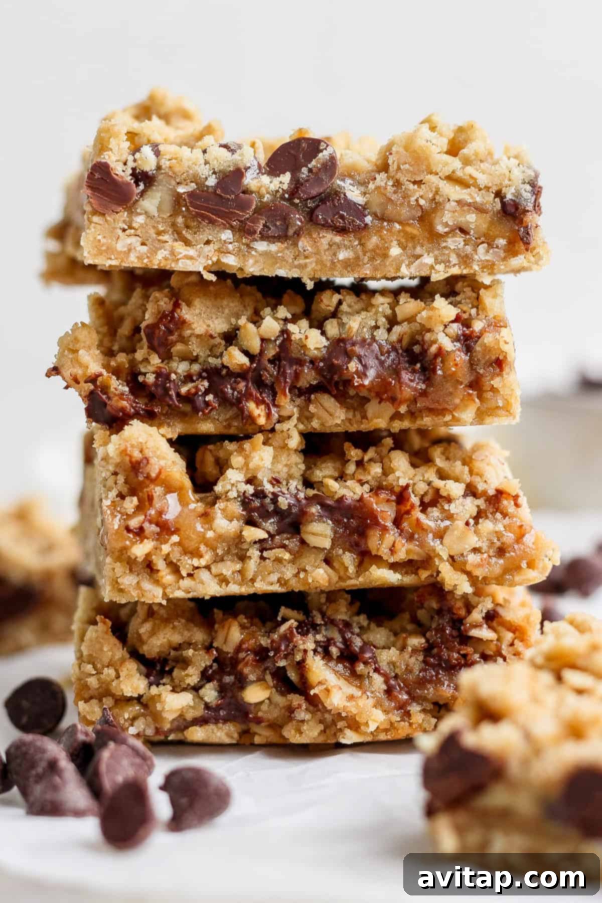 A stack of four chocolate chip oatmeal bars placed on a white surface, with scattered chocolate chips around them, highlighting their crumbly texture and delicious layers.
