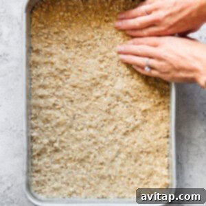 Hands gently pressing down a delicious oat-based mixture into a square baking pan. The mixture is evenly spread, forming the base of a dessert.